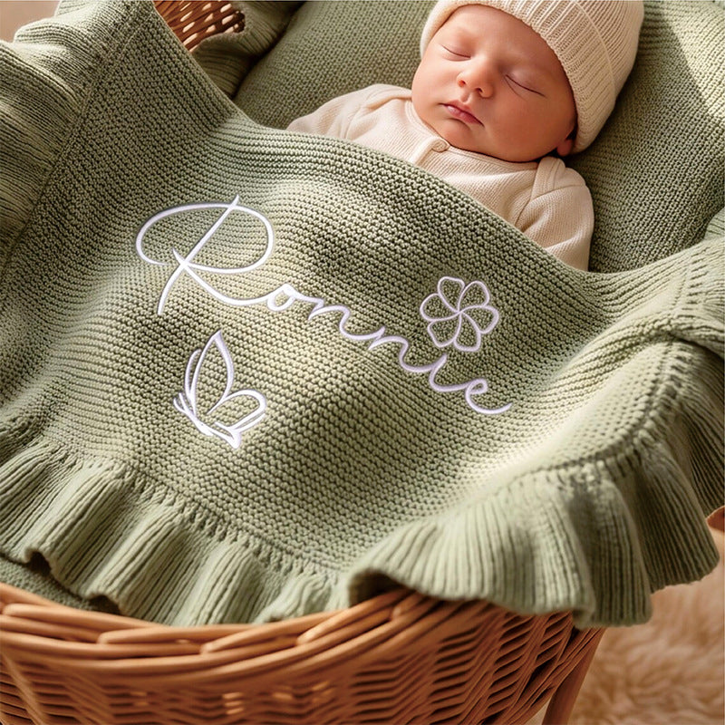 Newborn in a basket under a sage ruffle blanket embroidered with Ronnie