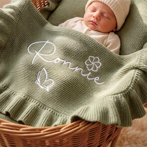 Newborn in a basket under a sage ruffle blanket embroidered with Ronnie