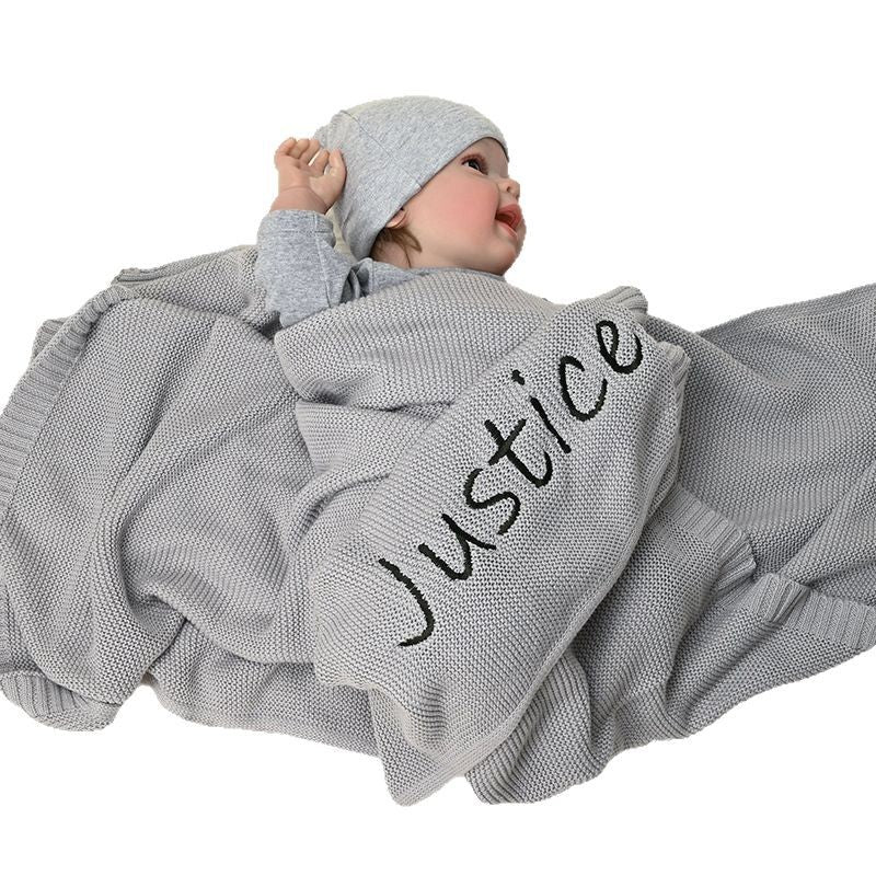 Baby under a gray knit blanket embroidered with Justice