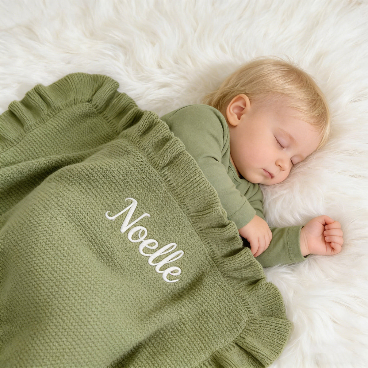 Baby asleep under a sage ruffle blanket embroidered with Noelle