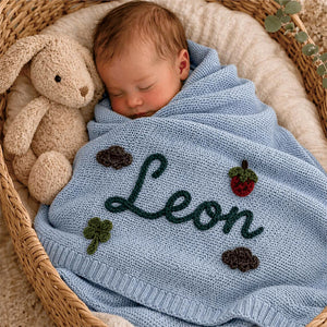 Newborn under a light blue knit blanket embroidered with Leon and motifs