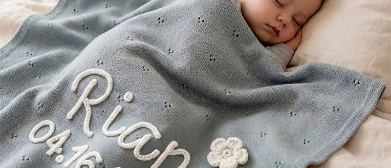Baby under a gray blue pointelle blanket embroidered with Rian and a date