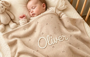 Baby asleep under a beige eyelet blanket embroidered with Oliver