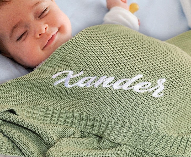 Smiling baby under a sage knit blanket embroidered with Xander