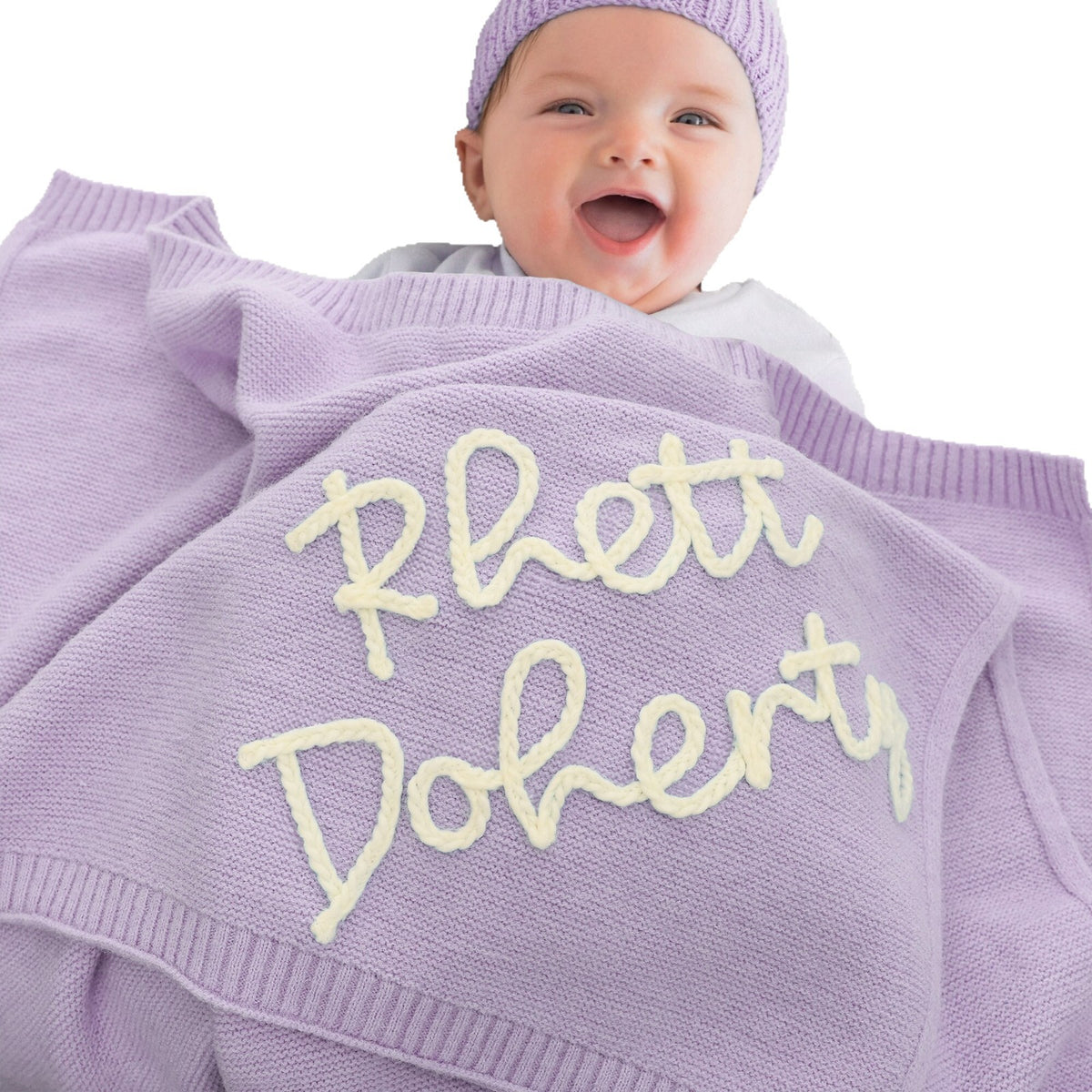 Smiling baby under a lavender blanket embroidered with Rhett Doherty