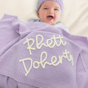 Smiling baby under a lavender blanket embroidered with Rhett Doherty