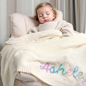 Sleeping toddler under a cream blanket embroidered with Ashley in rainbow yarn