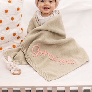Smiling baby under a beige blanket embroidered with Soraiya