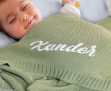 Smiling baby under a sage knit blanket embroidered with Xander